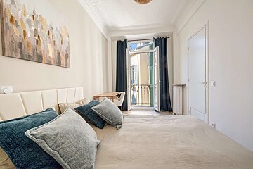 A bright, inviting bedroom featuring a comfy bed with blue and gray pillows, an abstract painting, and French doors opening to a balcony.