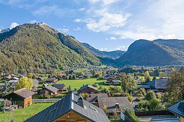 A picturesque mountain village nestled in a green valley, surrounded by lush, forested peaks under a bright blue sky.