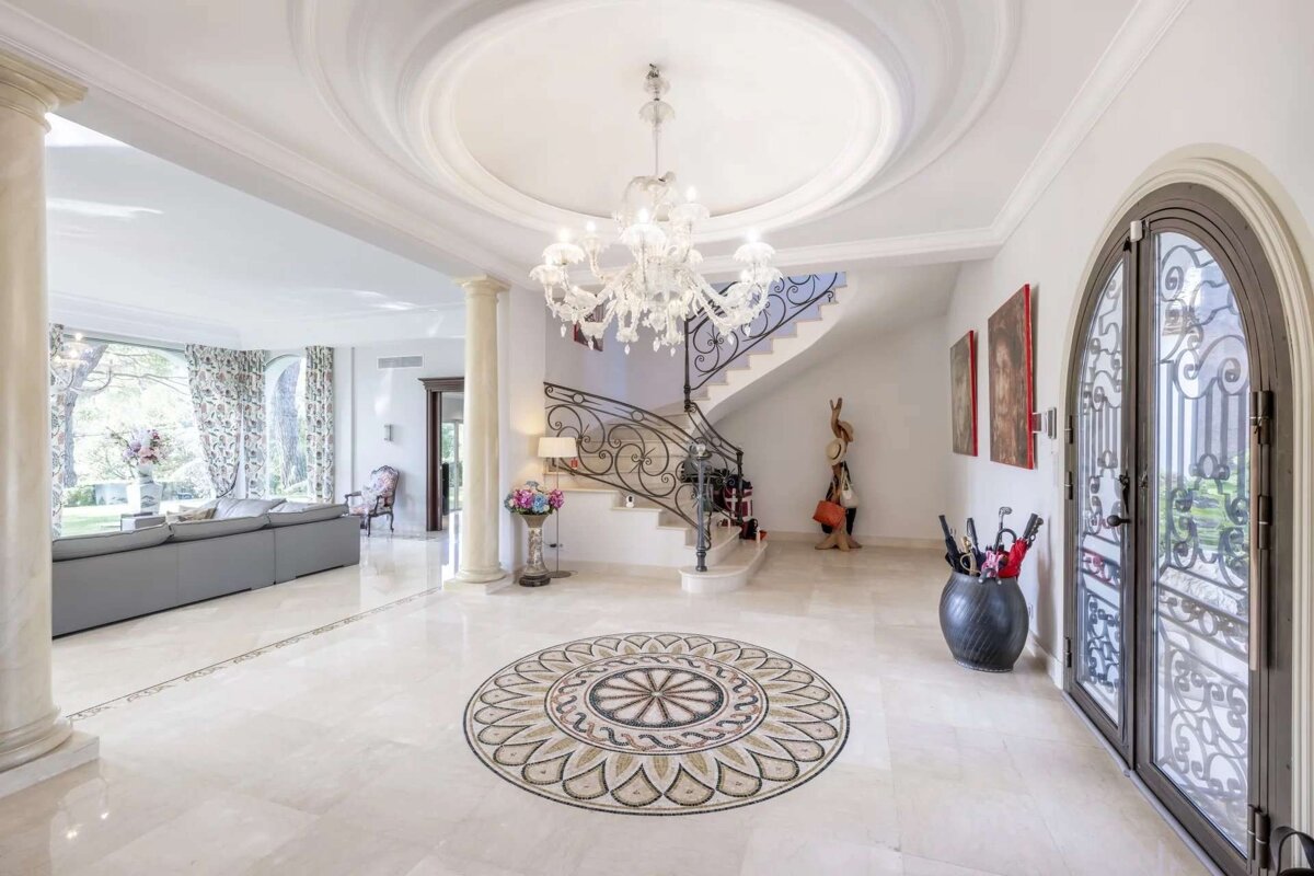 A luxurious, bright foyer featuring a grand chandelier, curved staircase, ornate floor mosaic, and elegant columns leading to a living area.