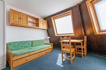 A rustic attic room with a green daybed, wooden wall cabinets, a small table & chairs, and bright windows on wood-paneled walls, all on a blue carpet.