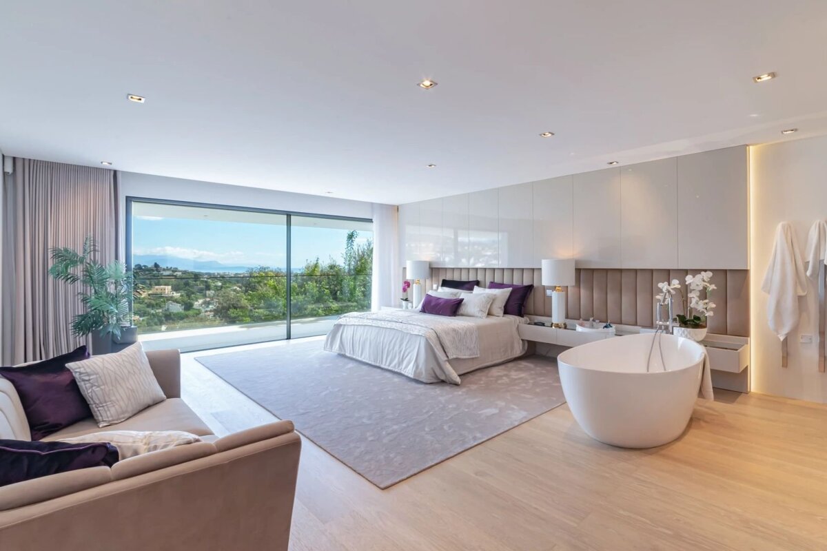 A luxurious modern bedroom with a large bed, an integrated bathtub, glass walls opening to a view, and purple accents.