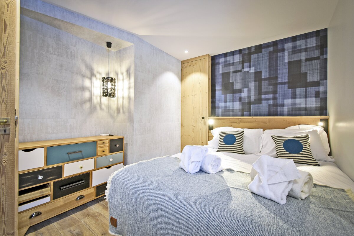 A modern bedroom featuring a bed with a blue blanket & towels, a geometric patterned headboard, and a unique multi-colored dresser.