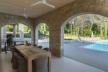 A spacious outdoor stone patio with an al fresco dining table, grill, and ceiling fans, opening via arches to a swimming pool and lush garden.