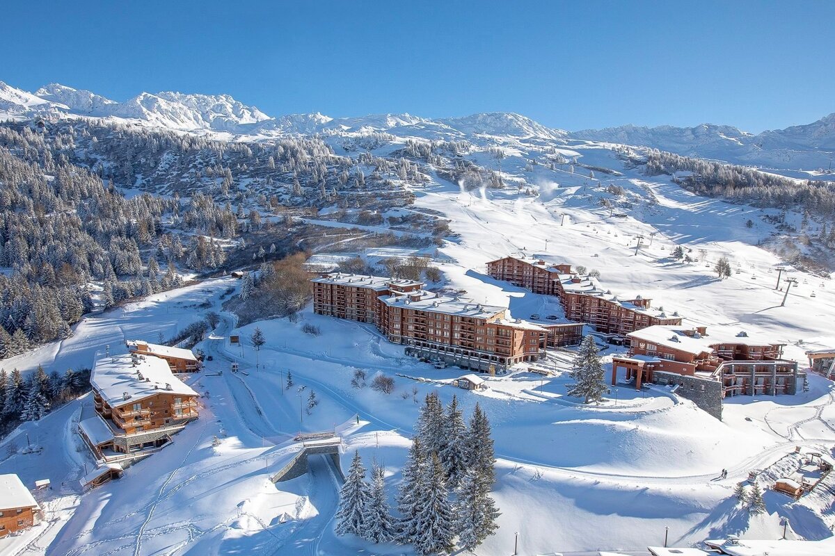 A sunny, snow-covered alpine resort with numerous lodges, vast ski slopes, and evergreen trees amidst majestic mountains under a clear blue sky.
