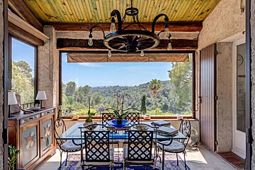 A rustic dining room with a glass table, wrought iron chairs, and a large window offering panoramic views of a green, hilly forest.