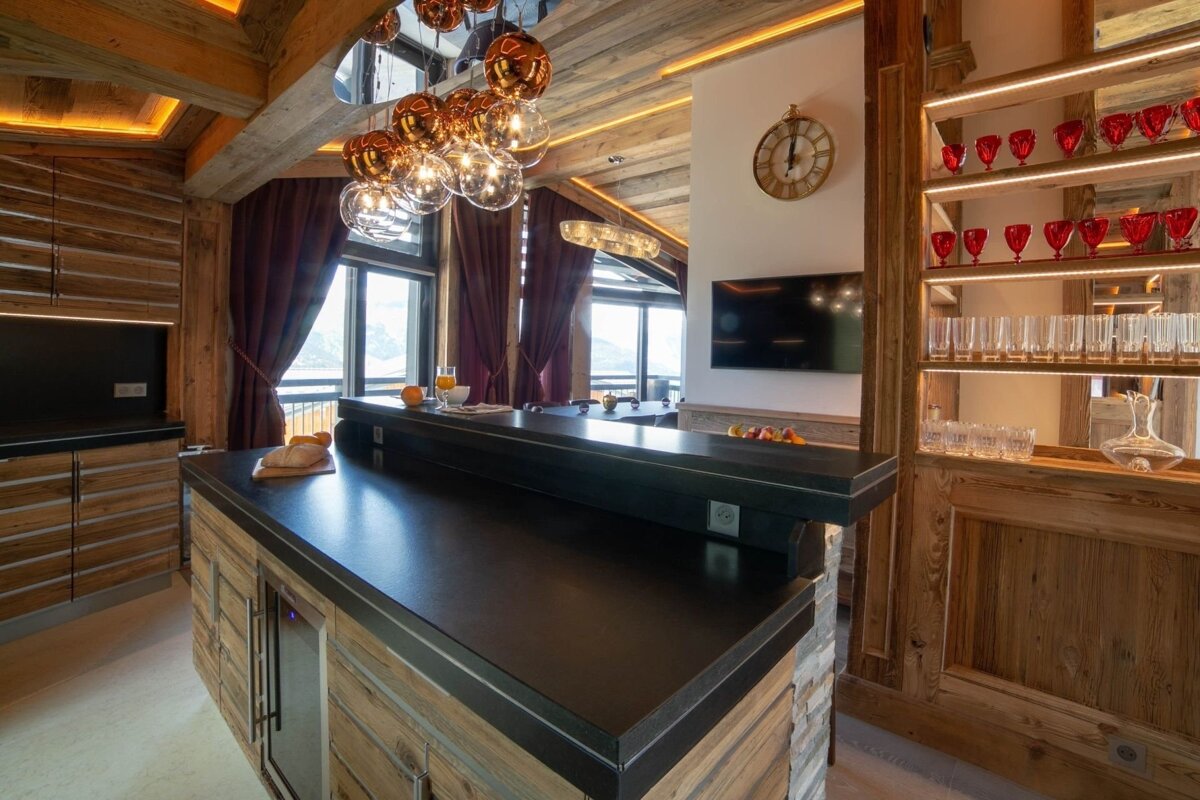 Elegant, modern chalet kitchen and bar with wooden beams, dark countertops, stylish lighting, and stunning mountain views from large windows.