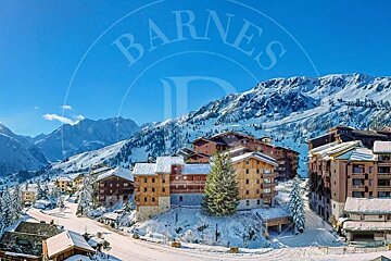 Snow-covered wooden chalets in a mountain ski resort under a clear blue sky, featuring a BARNES watermark.