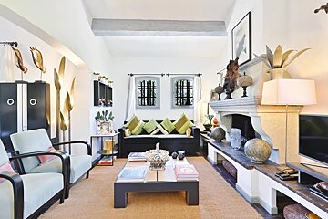 An elegant, rustic living room featuring a large stone fireplace, black sofa with green pillows, light armchairs, golden decor, and a white ceiling beam.