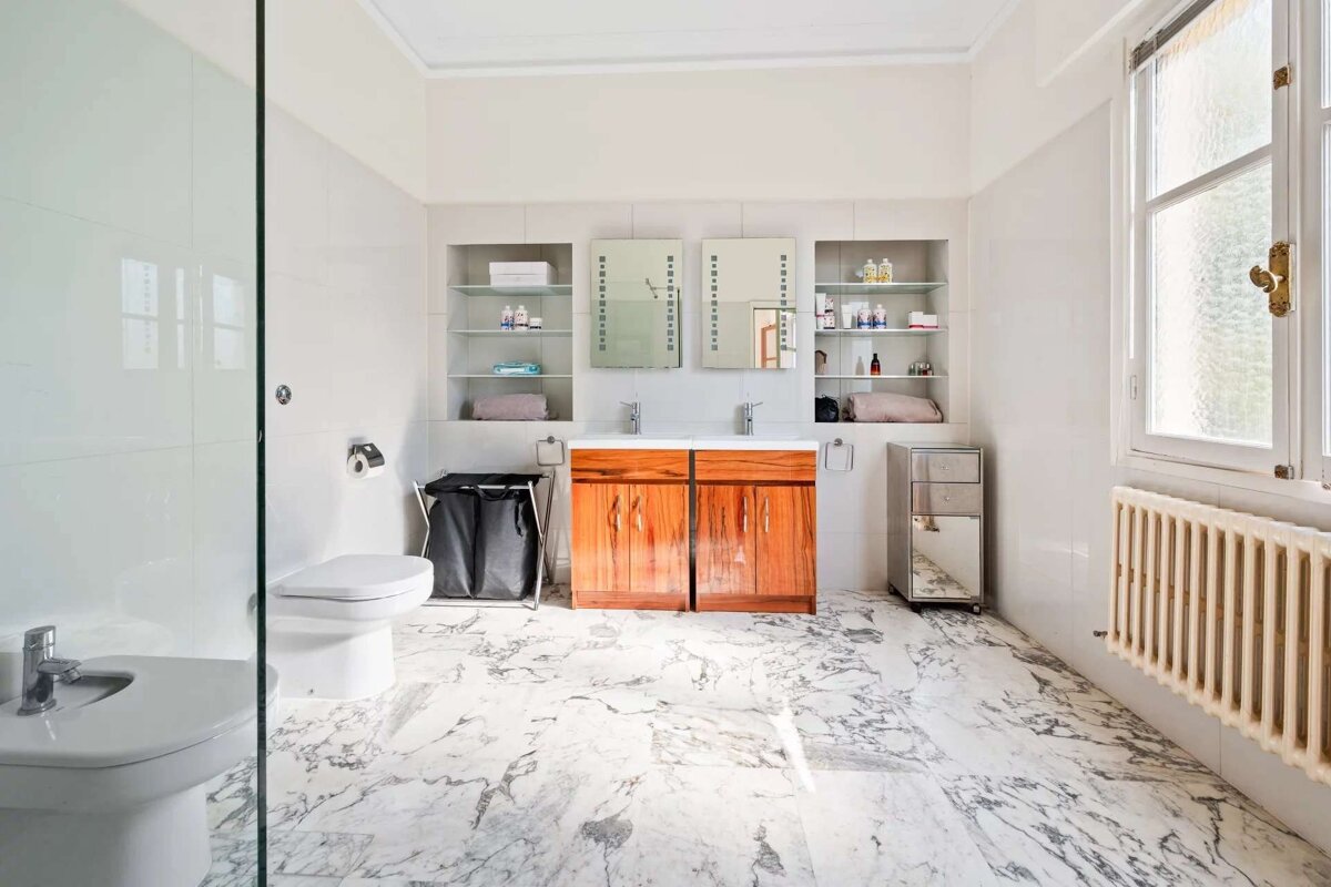 Bright, spacious bathroom with white marble floors, tiled walls, double wooden vanity, two mirrors, toilet, bidet, and a large window.