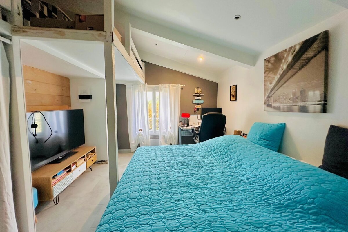 A bright bedroom with a blue quilted bedspread, a desk area, TV console, and a visible loft bed structure, featuring a sloped ceiling.