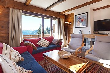 Cozy cabin living room with red sofas, a wooden coffee table, grey armchairs, and a large window offering a scenic mountain view.