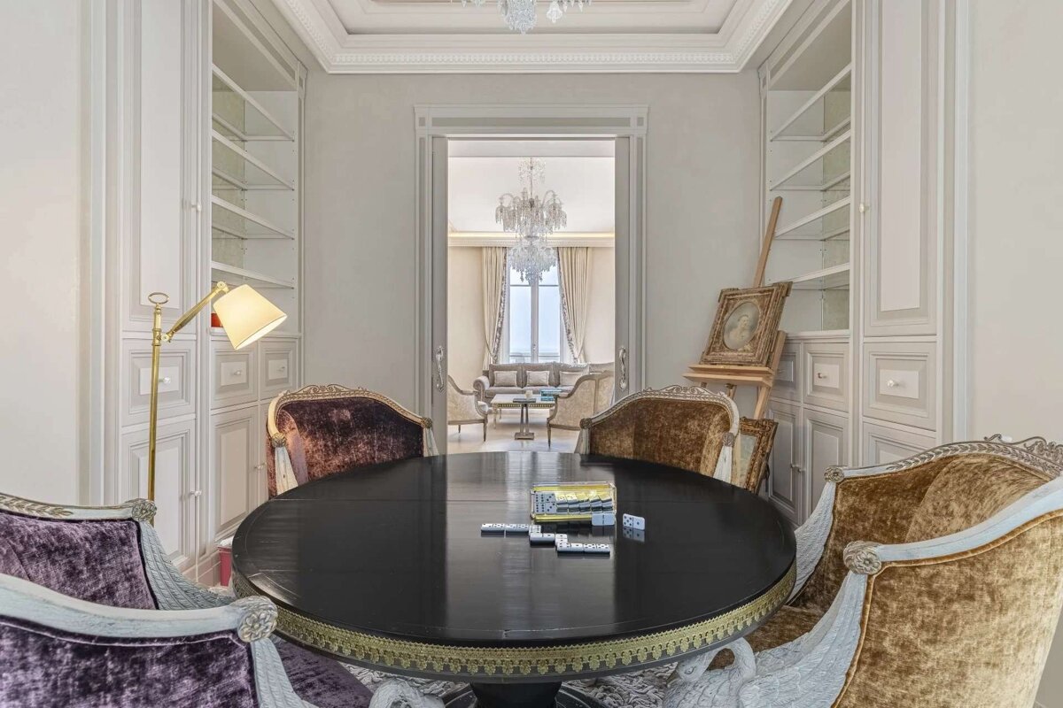 An elegant room features a dark oval table with ornate velvet chairs and dominoes. Built-in bookcases flank a doorway leading to a luxurious living area.