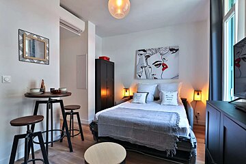 Modern studio apartment with a bed, a high-top table with stools, and pop art on the wall. Features light wood floors and minimalist decor.