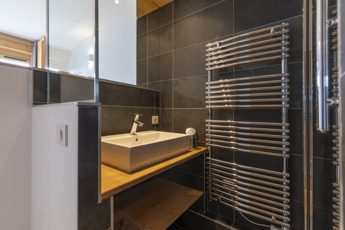 Modern bathroom with black tiles, white vessel sink on wood vanity, chrome towel warmer, and a glass partition.