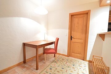 A brightly lit room with a wooden desk, red chair, and lamp next to a wooden door. A floral rug covers a tiled floor.