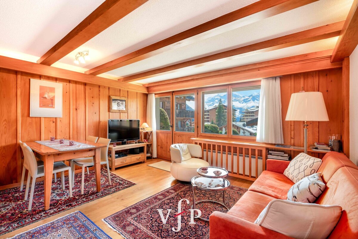 Cozy wood-paneled chalet living room featuring an orange sofa, dining area, and large windows with scenic mountain views.