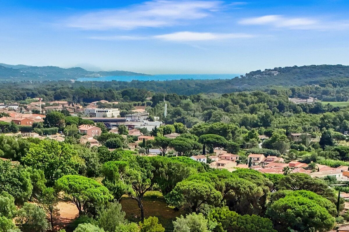 High-angle view of a coastal town surrounded by lush green forests and hills under a blue sky, with the ocean in the distance.