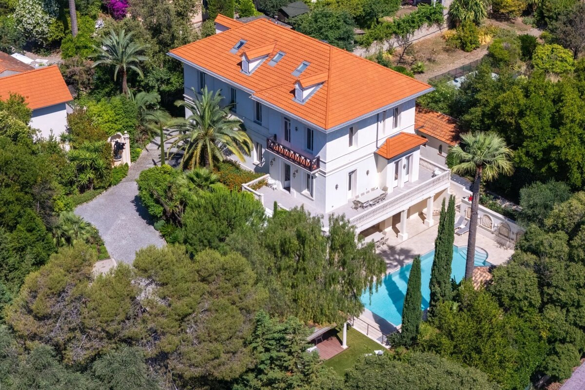 Aerial view of a grand white villa with a vibrant orange roof, surrounded by lush green trees, palm trees, a swimming pool, and stone paths.