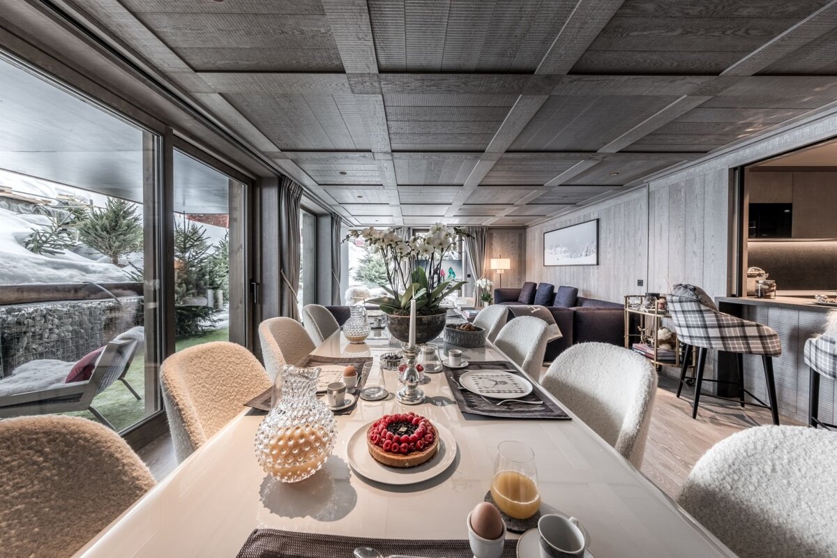 Chic chalet dining room with a breakfast-laden table, plush seating, and large windows overlooking a snowy outdoor scene.
