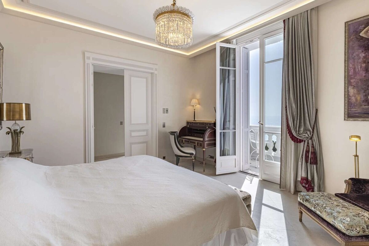A luxurious bedroom featuring a large bed, an elegant writing desk, and a balcony with an ocean view. Natural light fills the neutral-toned room.