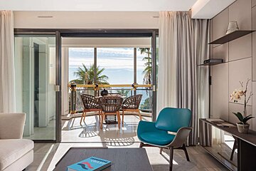 A modern living room with a blue armchair and a balcony offering stunning sea views, complete with palm trees and a stylish outdoor table setting.