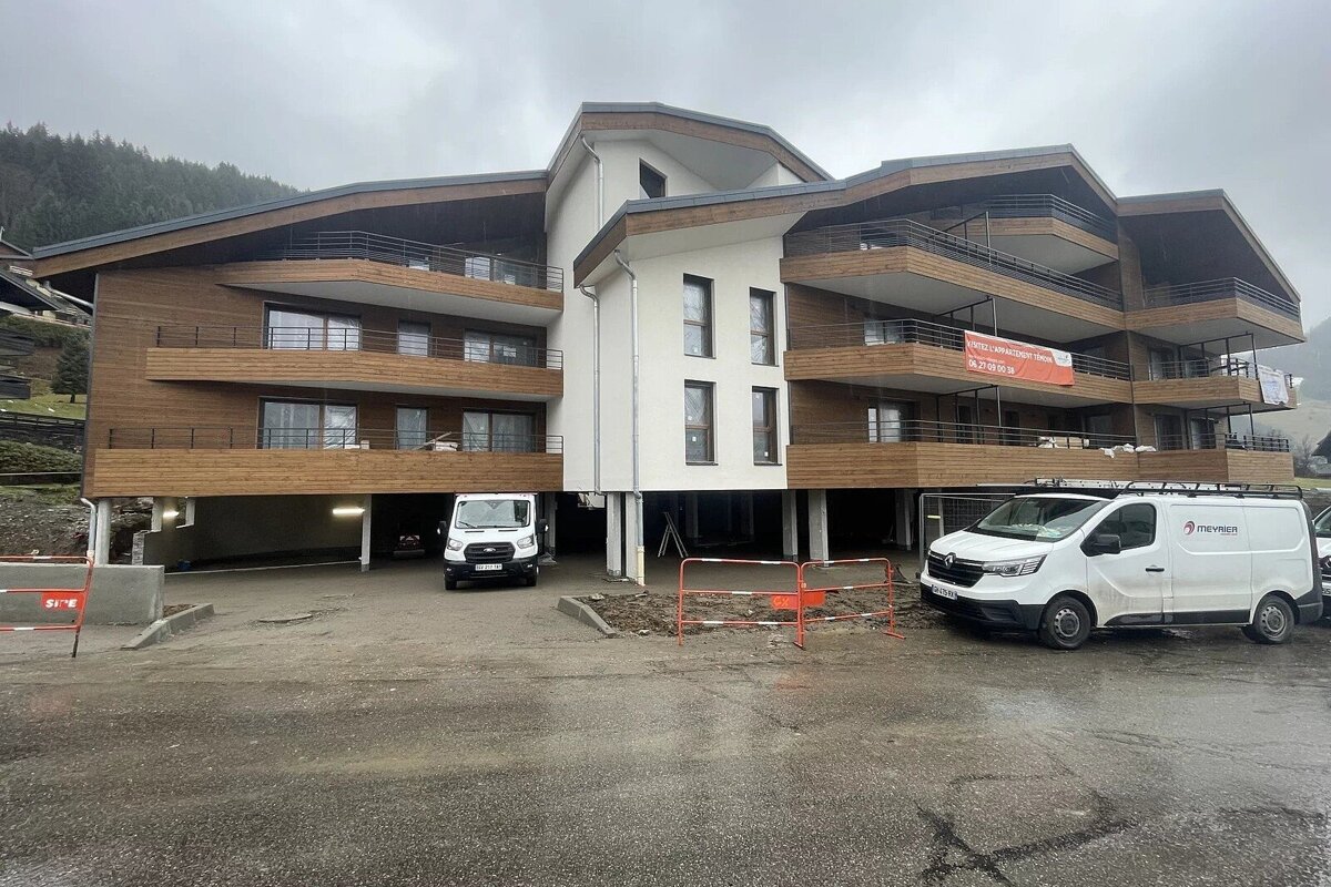 Modern wood and white apartment building with balconies, under construction, with two white vans parked in front on an overcast day.