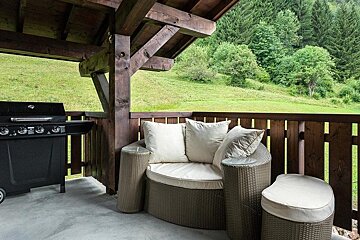 A covered wooden patio featuring a black gas grill, comfortable wicker seating, and a scenic view of green hills and forests.