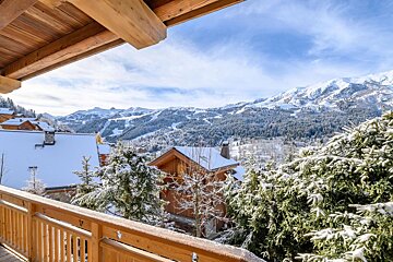 View from a wooden balcony overlooking a snowy mountain village with chalets, snow-covered trees, and distant ski slopes under a blue sky.