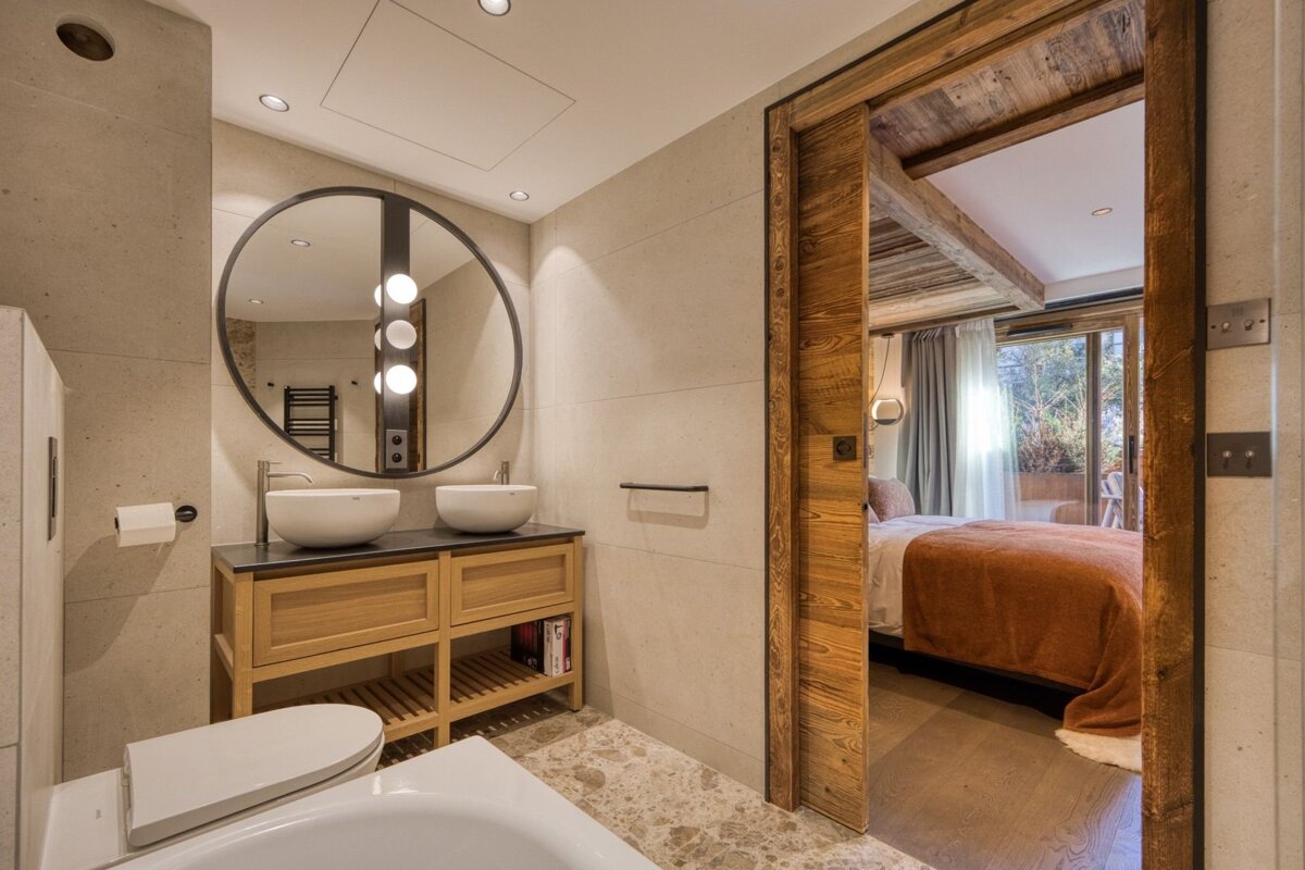 Modern bathroom with double vanity, round mirror, and a wooden sliding door leading to a cozy bedroom with a balcony.