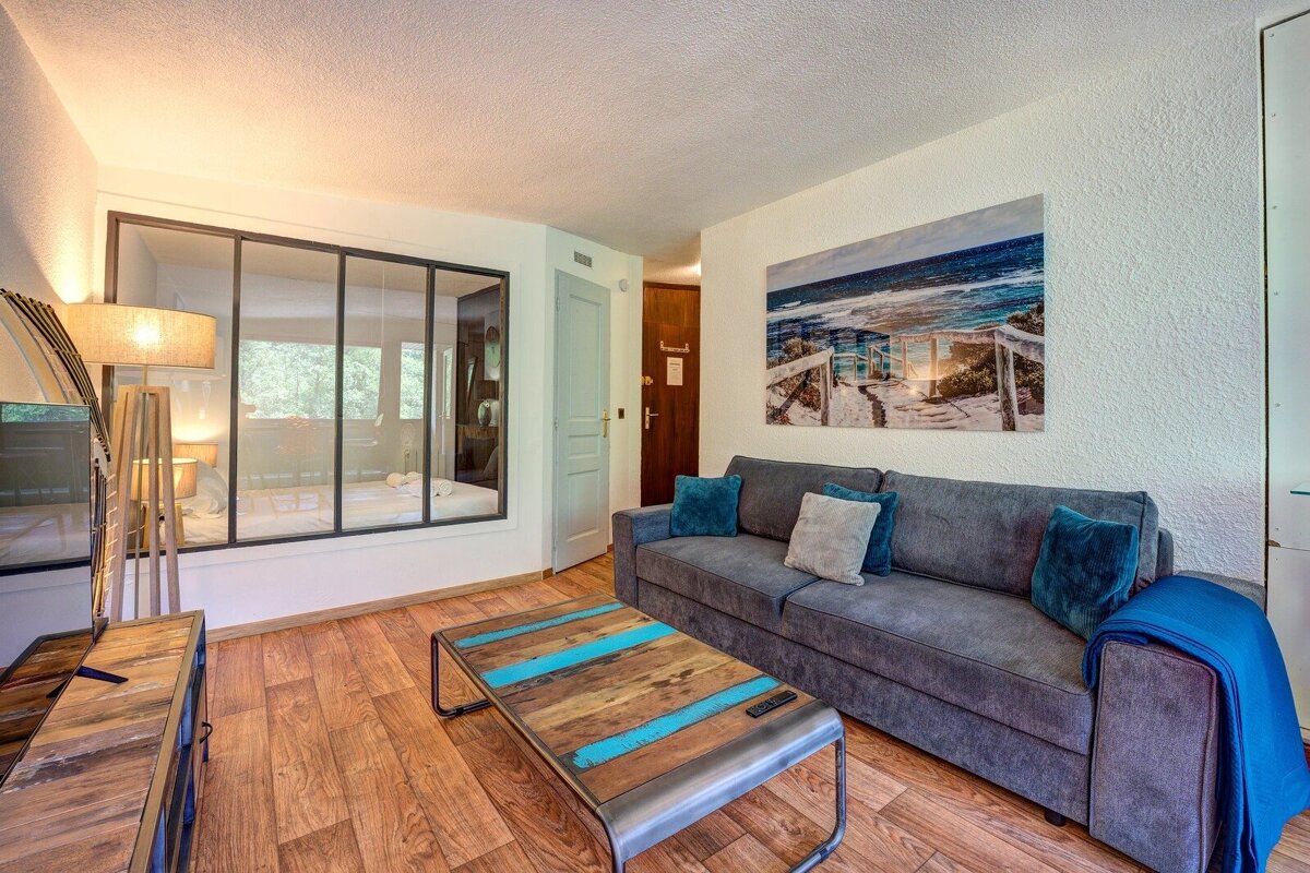 Cozy modern living room featuring a grey sofa, rustic coffee table, beach wall art, and a glass-partitioned bedroom.