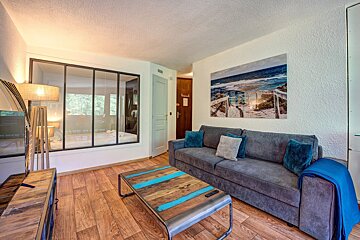 Cozy modern living room featuring a grey sofa, rustic coffee table, beach wall art, and a glass-partitioned bedroom.