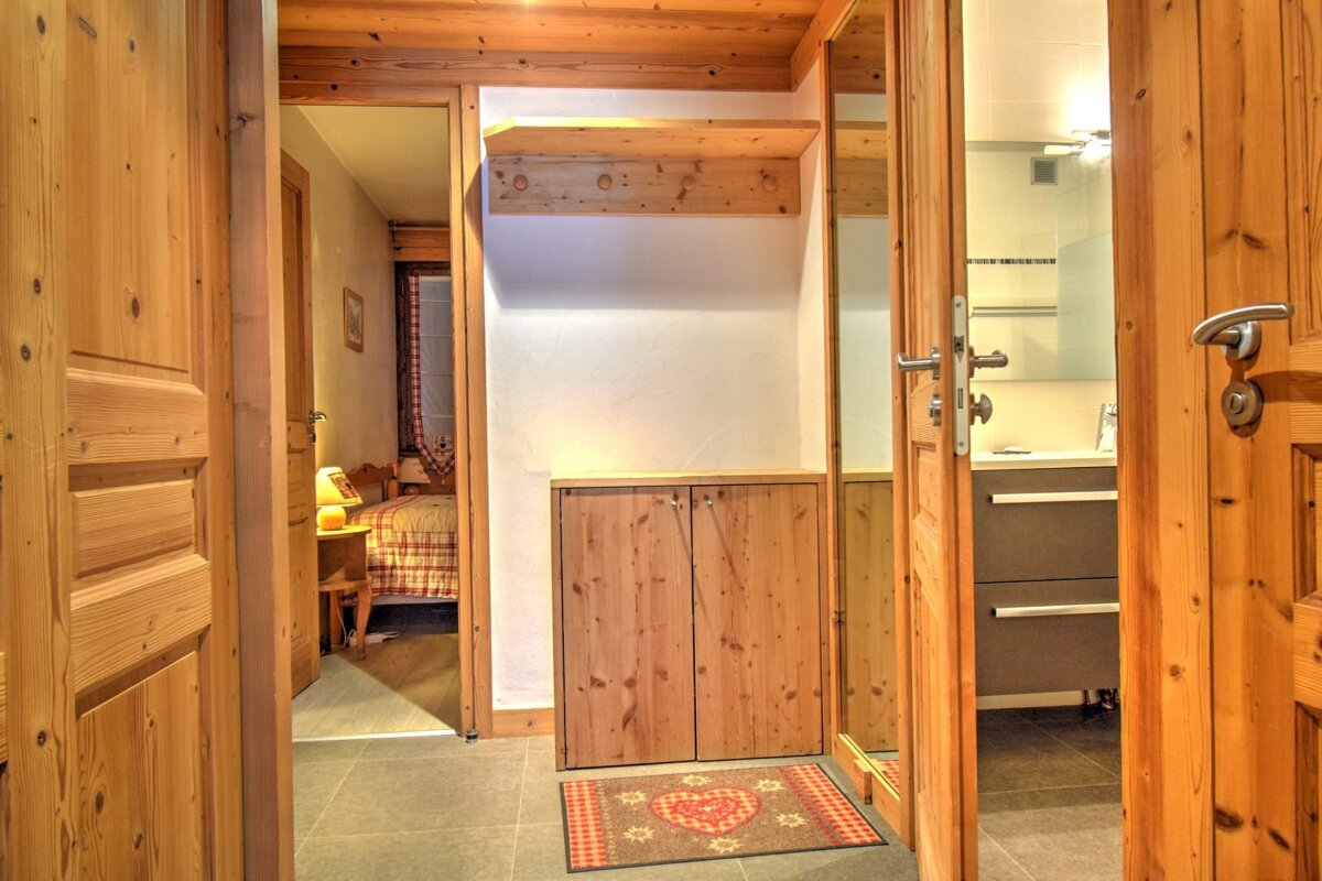 Rustic wooden hallway featuring open doors to a cozy bedroom (left) and a modern bathroom (right). A central entryway has a coat rack, cabinet, and heart-patterned mat.