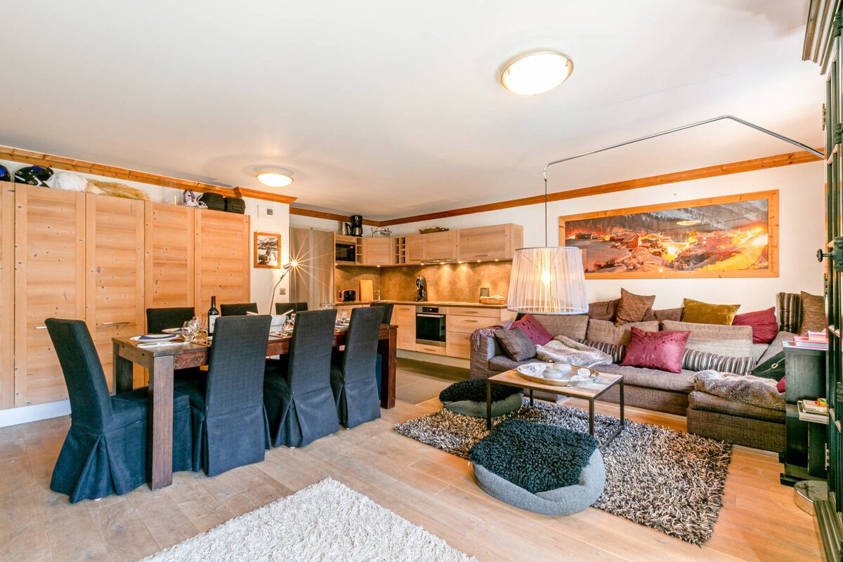 A bright, open-plan living area with a dining table, comfy sectional sofa, modern kitchenette, and warm wooden furnishings.