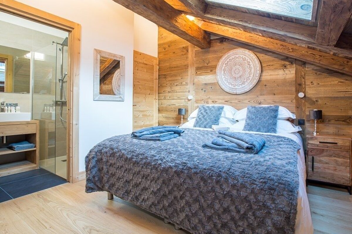 Cozy chalet bedroom featuring wood-paneled walls, exposed ceiling beams, a plush grey bed, and a peek into the ensuite bathroom.