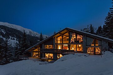 A cozy, brightly lit chalet nestled in a snow-covered mountain landscape at dusk, with warm lights glowing from its many windows.