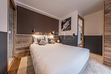 A modern, cozy bedroom with a large bed, rustic wooden headboard, brown and grey walls, and black-and-white art. Features warm wood accents.