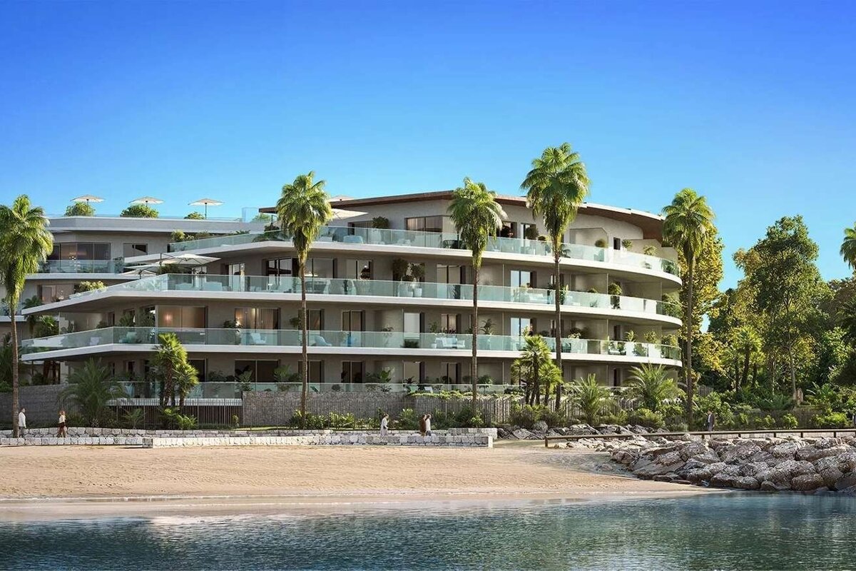 Modern waterfront apartment complex with tiered balconies, palm trees, sandy beach, and clear water under a bright blue sky.
