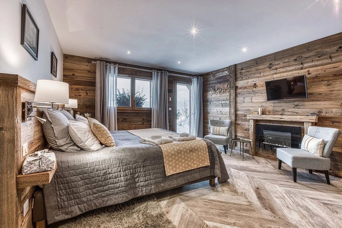 Luxurious chalet bedroom with rustic wood paneling, a comfy bed, fireplace, and TV. Windows reveal a snowy mountain view, creating a warm, cozy retreat.