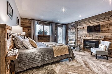Luxurious chalet bedroom with rustic wood paneling, a comfy bed, fireplace, and TV. Windows reveal a snowy mountain view, creating a warm, cozy retreat.