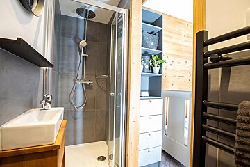 Modern bathroom featuring a glass shower, vessel sink on a wooden vanity, grey tiled walls, blue shelving with plants, and a black towel warmer.