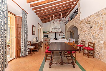 A rustic, open-plan room with a dining table, living area with a fireplace, exposed wooden beams, and a stone wall with an archway.