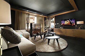 Chic, dark-themed living room with wood accents, a cozy sofa, nested coffee tables on a round rug, and a TV playing 20th Century Fox.