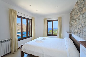 A bright bedroom with a white bed, two windows overlooking a blue sea and rocky coastline, and a textured stone accent wall.