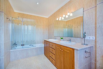 A spacious, light brown tiled bathroom featuring a bathtub with a glass shower screen, a double wooden vanity, and a large mirror with lights.