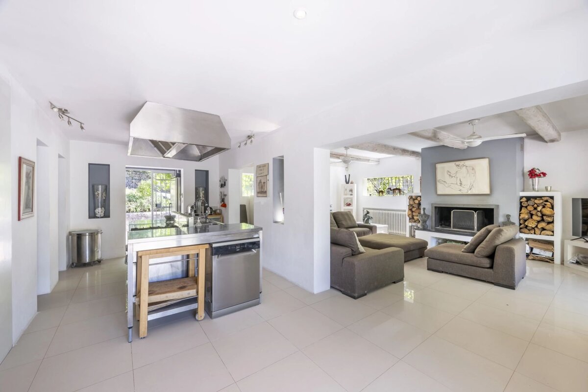 A modern, open-plan white interior featuring a stainless steel kitchen island next to a living room with grey sofas and a fireplace.