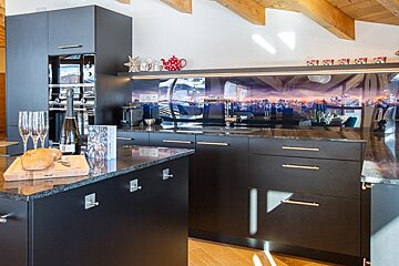 Modern black kitchen featuring a scenic mountain backsplash, wooden ceiling beams, and an island with bread and champagne.