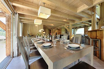 A cozy, upscale dining area with a long, set wooden table, featuring exposed timber beams, plank walls, and modern square pendant lights.