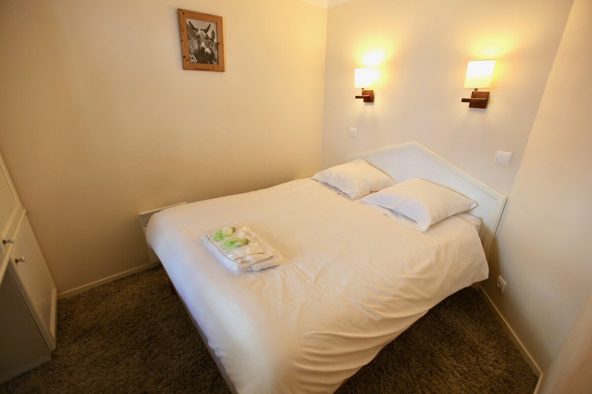 A compact, brightly lit bedroom with a double bed, white sheets, two wall lights, a picture of donkeys, and brown carpet. Fresh towels await on the bed.