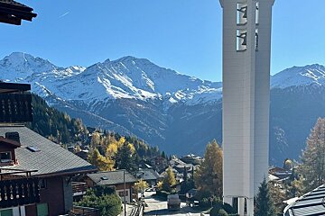 Alpine village view with a tall white bell tower in the foreground and majestic snow-capped mountains in the background.