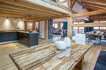 Modern rustic chalet interior featuring a glass-topped wooden table, open kitchen, and exposed wooden beams throughout.